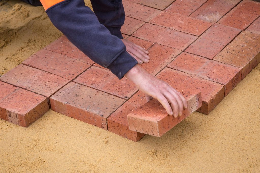 Worker's hand laying red bricks on sand for pavement construction.